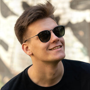 A man wearing SUL EXTRAS Visi sunglasses with shiny black rectangular frames and green lenses outdoors.
