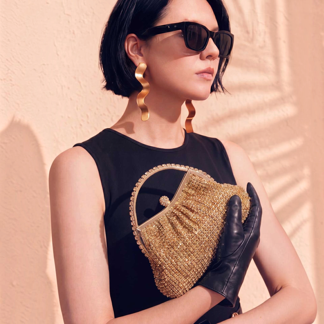 SUL EXTRAS Estelle evening bag in gold rhinestone with arched handle, held by a woman in black dress and gloves.