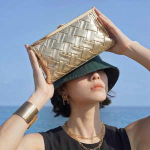 SUL EXTRAS Solène metallic gold woven evening clutch held by a model wearing a dark green bucket hat by the sea.