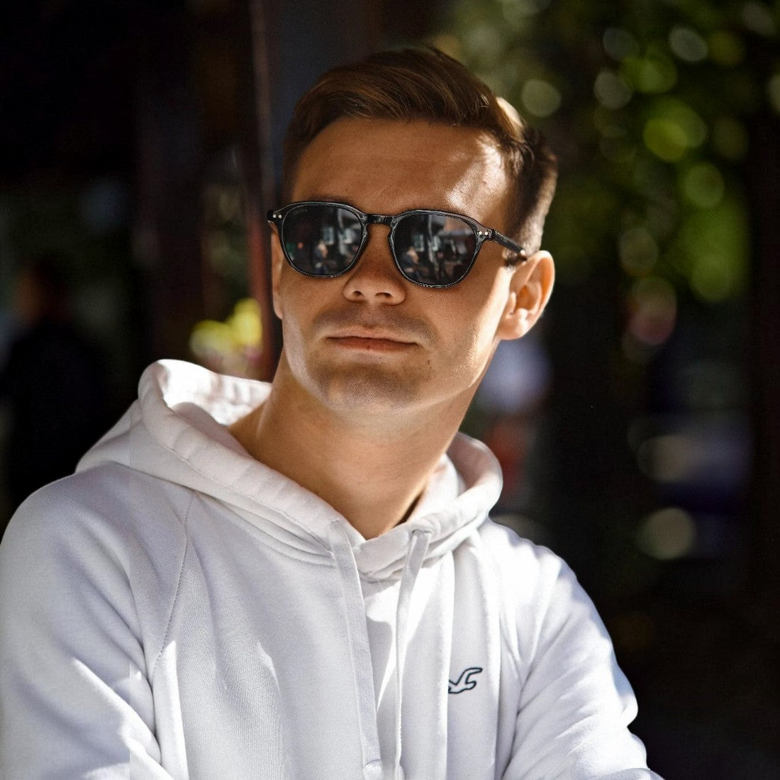 Man wearing SUL EXTRAS Ace sunglasses with black frames and dark lenses, styled with a white hoodie outdoors.