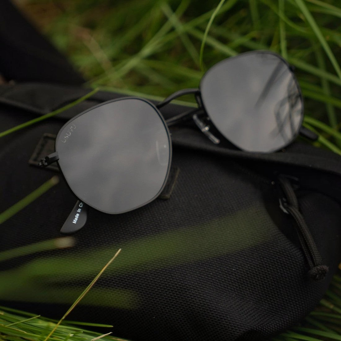 SUL EXTRAS Eternally minimalist geometric sunglasses with black metal frames and dark lenses resting on a black bag