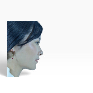 A model wearing SUL EXTRAS gold-plated sterling silver drop earrings with a pearl and red zirconia stone.