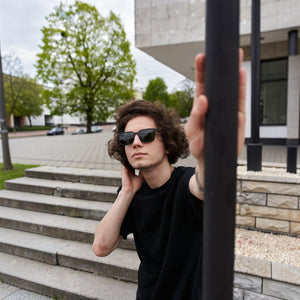 A young man with curly hair wearing SUL EXTRAS Leo polarized sunglasses with a black frame outdoors.