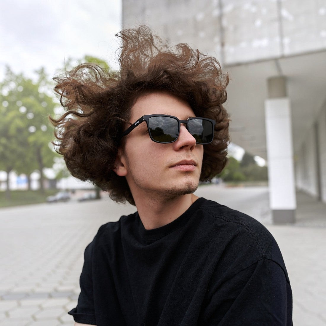 A young man with curly hair wearing SUL EXTRAS Leo polarized sunglasses with a black frame outdoors.