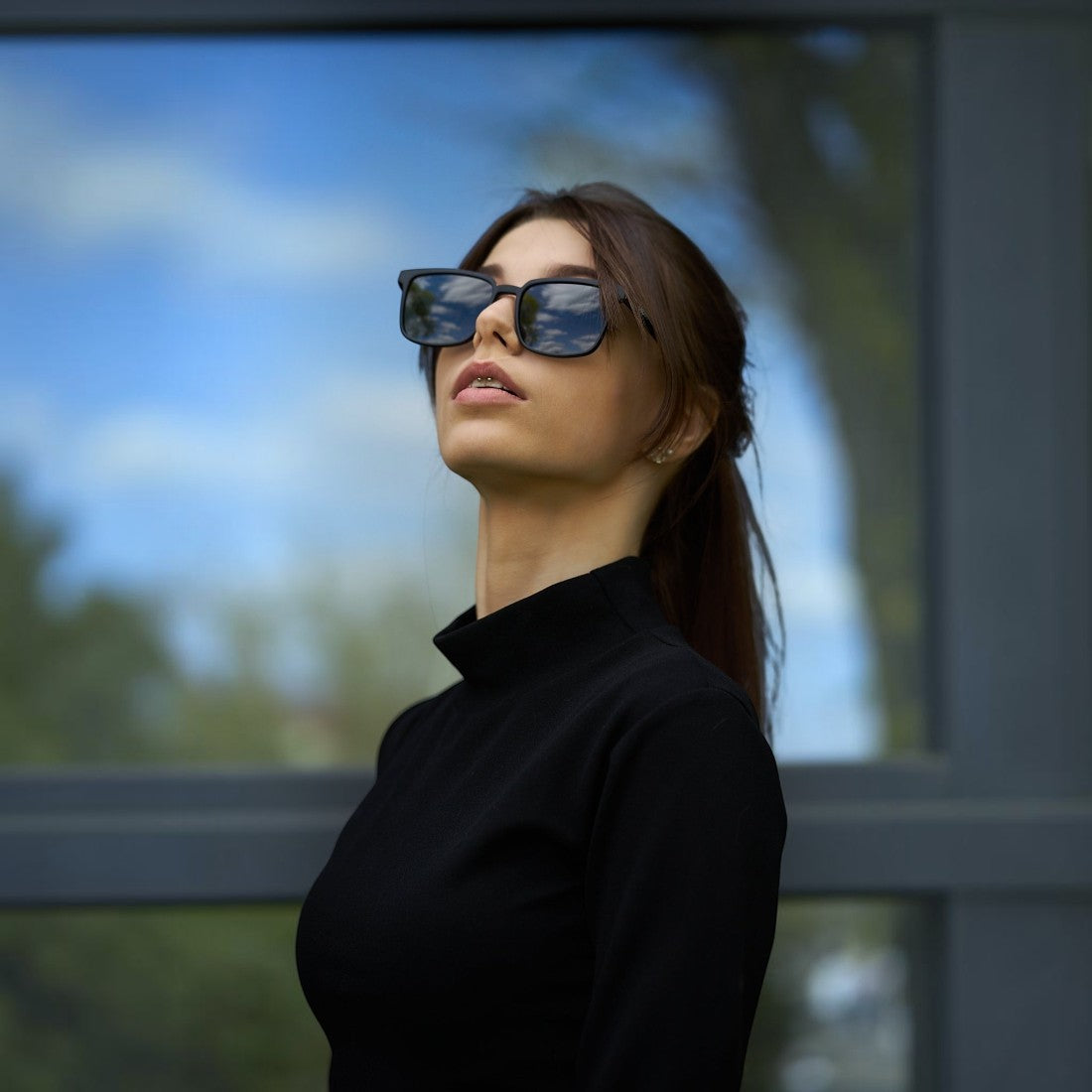 Woman wearing SUL EXTRAS Leo polarized sunglasses with black frames and dark lenses against a glass building background