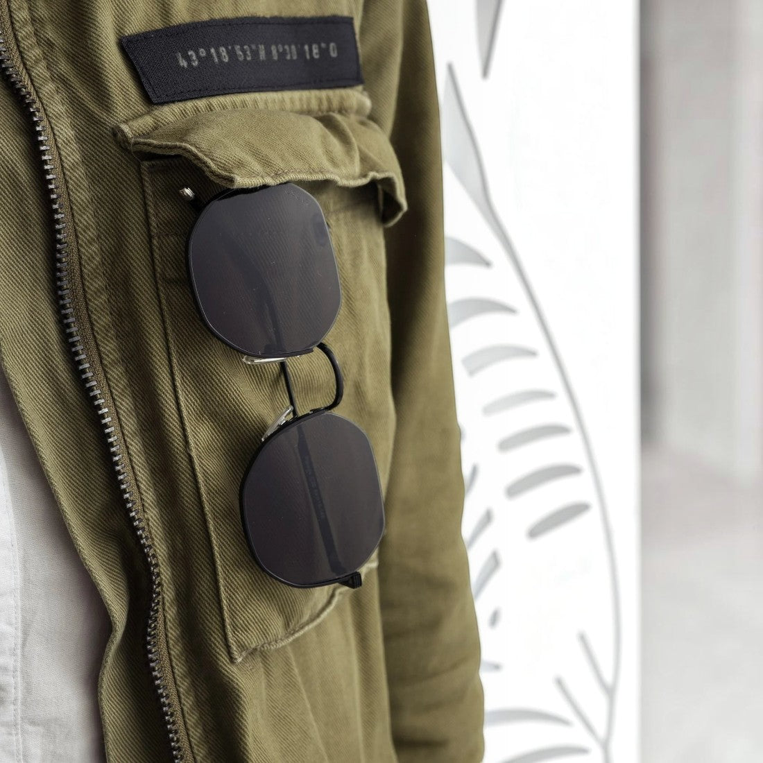 SUL EXTRAS Parade sunglasses with black geometric round frames and smoke lenses hanging from an olive green jacket