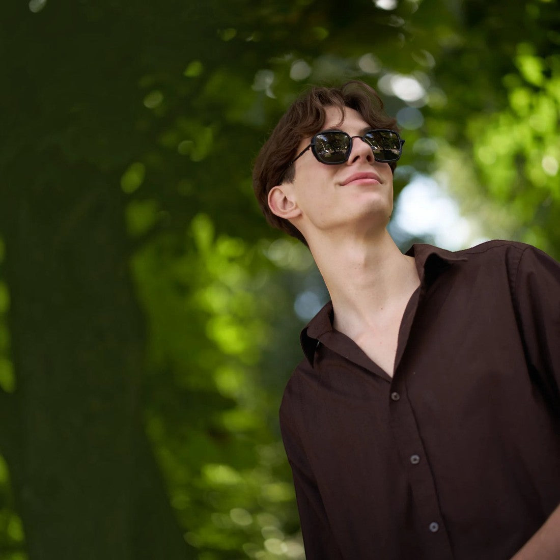 A young man wearing SUL EXTRAS Spark polarized sunglasses with a durable black frame outdoors in a park setting.