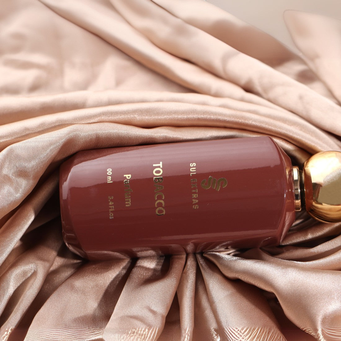 SUL EXTRAS Tobacco perfume bottle in a deep brown glass with gold lettering, resting on elegant champagne silk fabric.