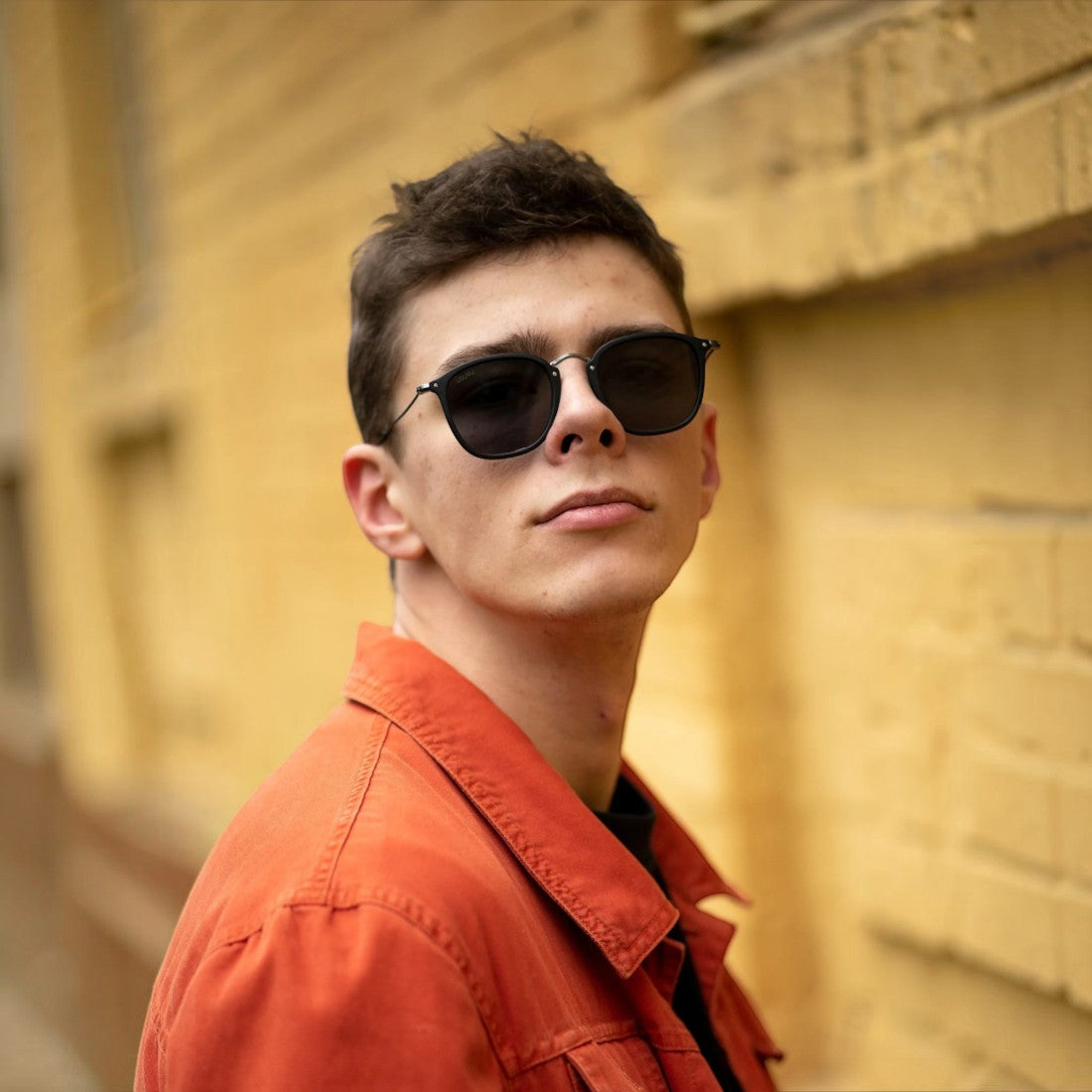 SUL EXTRAS Visi sunglasses with shiny black rectangle frames and green lenses worn by a man in an orange jacket